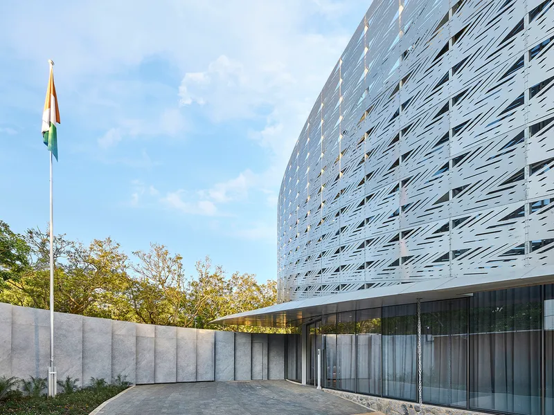 Ground-level view of the perforated metal facade meeting a concrete wall and glazed entrance below afternoon clouds