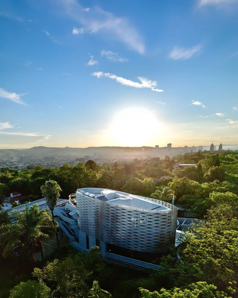 Drone view of the oval building with white screen facade surrounded by trees at sunset