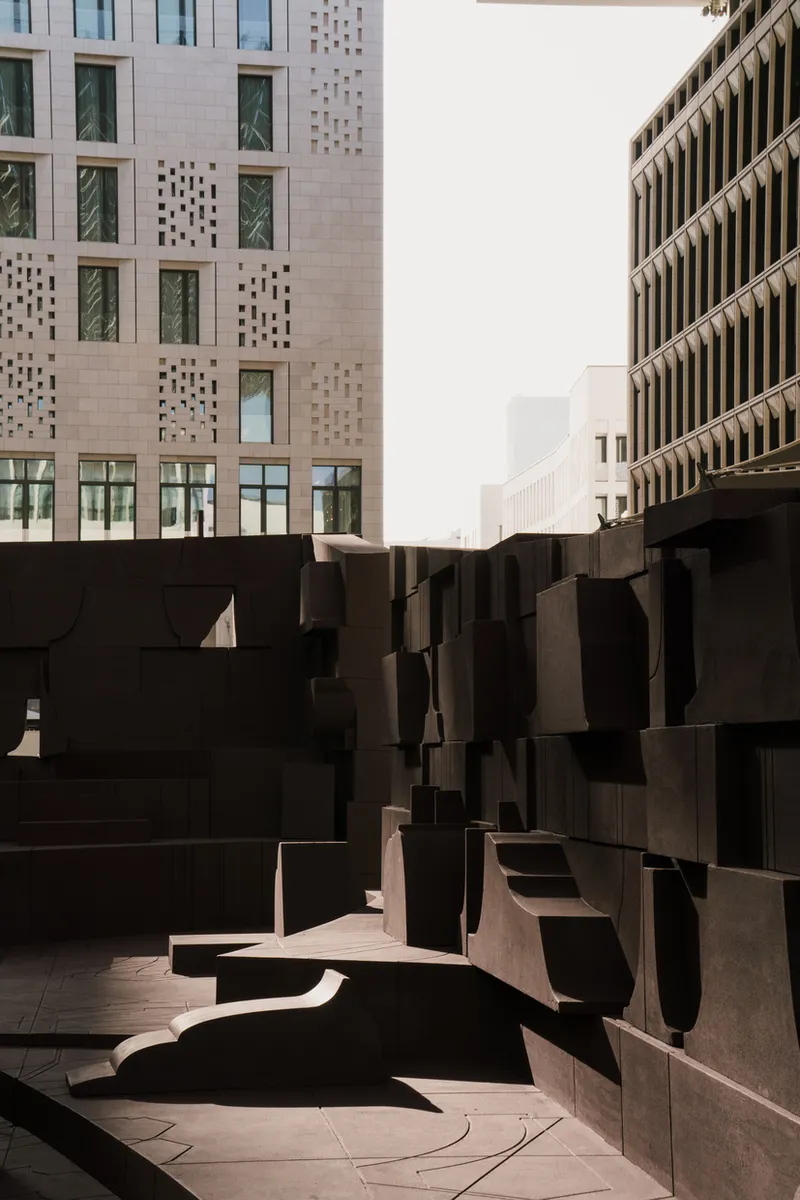 Narrow passage between dark sculpted seating blocks with perforated facade visible beyond in hazy daylight