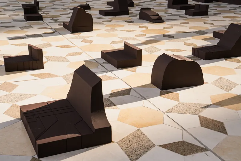 Geometric paving pattern of stone and terrazzo tiles with dark bronze sculptural seating forms in daylight
