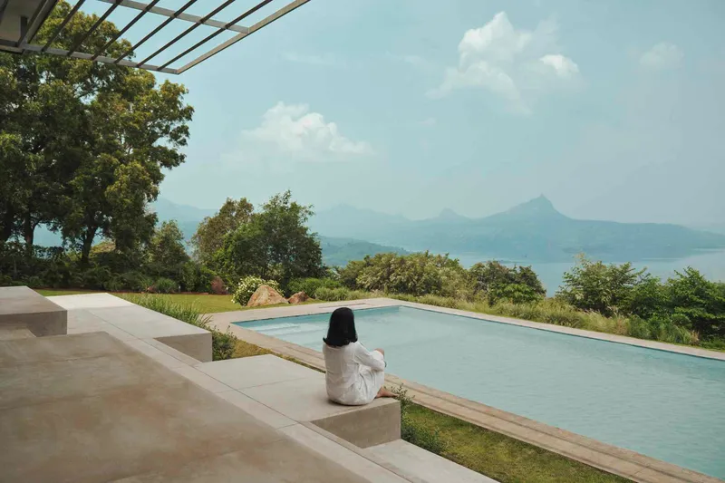 Infinity pool terrace with concrete seating ledge where a person sits overlooking mountains and lake