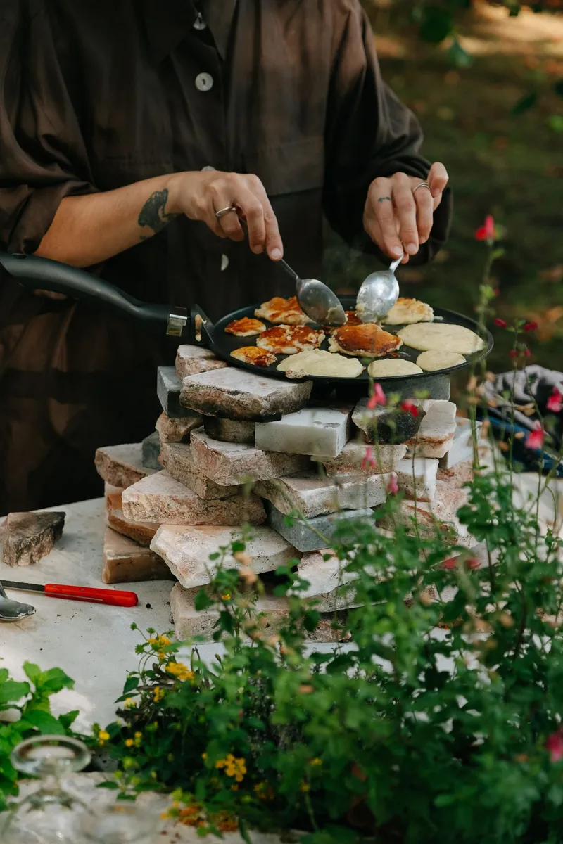 Hands working a flat griddle atop a stacked stone cooking platform surrounded by flowers