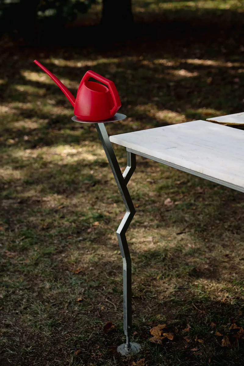 Red watering can resting on the zigzagging steel support of an outdoor table in dappled sunlight