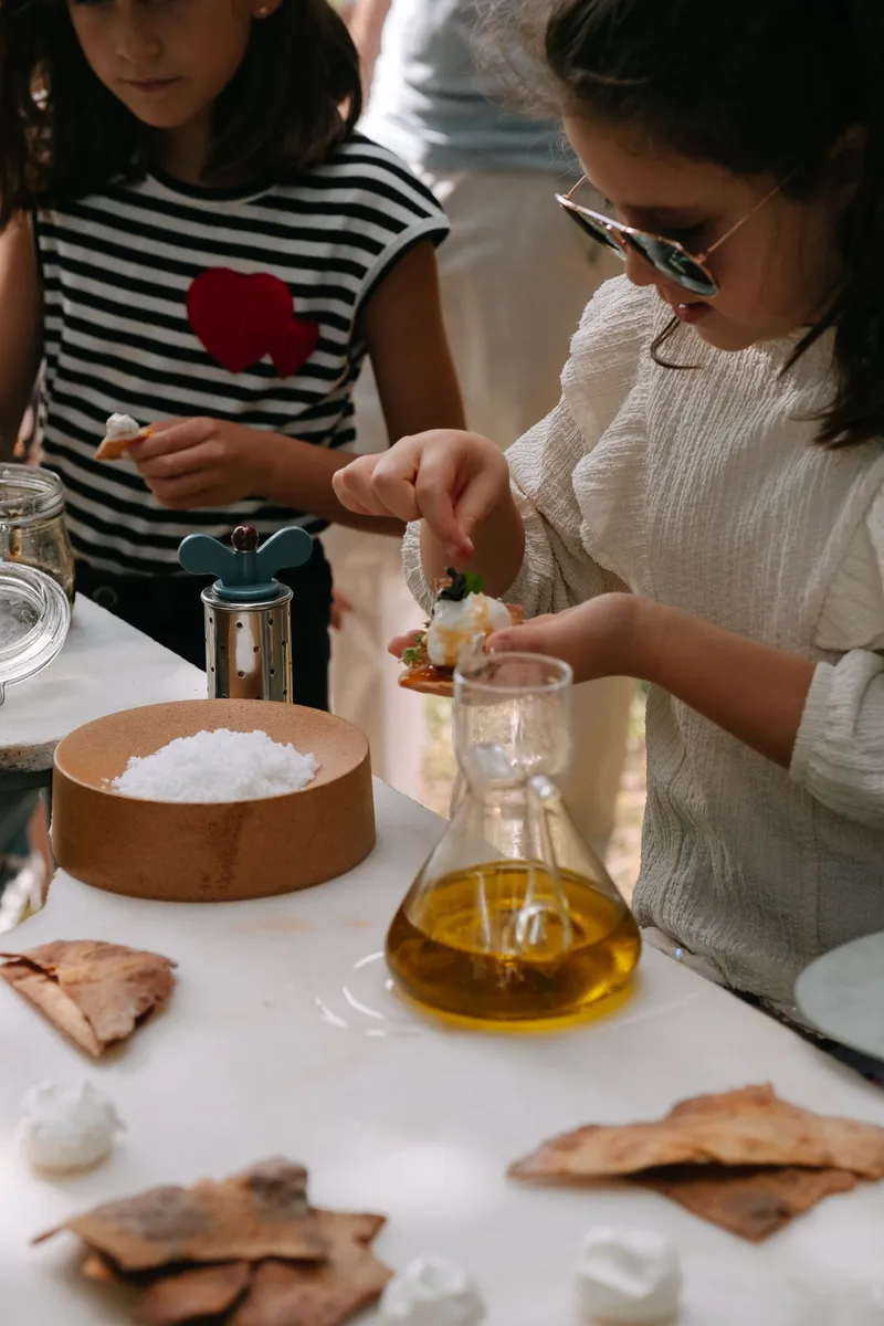 Two children preparing food at a white table with olive oil, salt, and flatbread