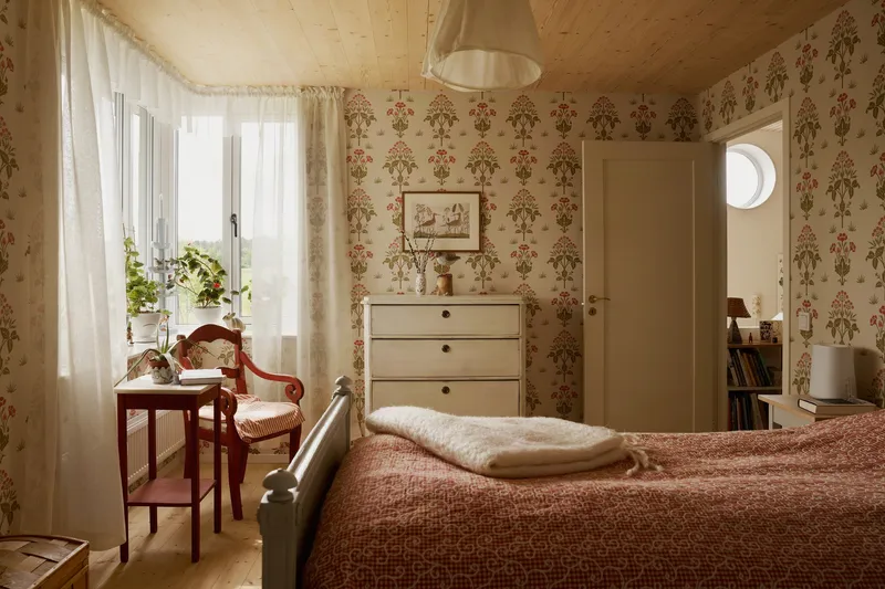 Bedroom with floral patterned wallpaper, exposed timber ceiling and sheer curtains filtering daylight