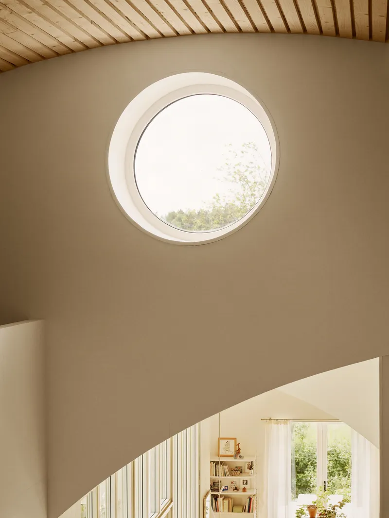 Circular window in a taupe wall beneath a timber slat ceiling with daylight filtering through
