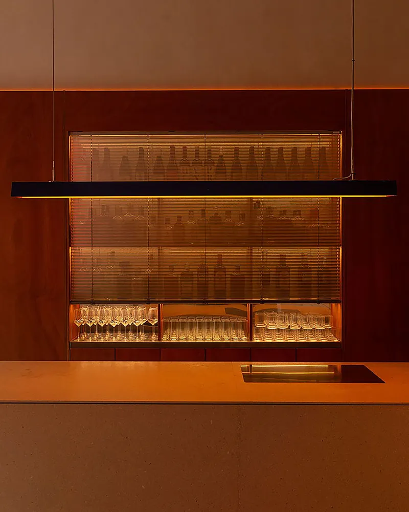 Backlit bar display cabinet with glassware behind a suspended linear light fixture and terrazzo counter