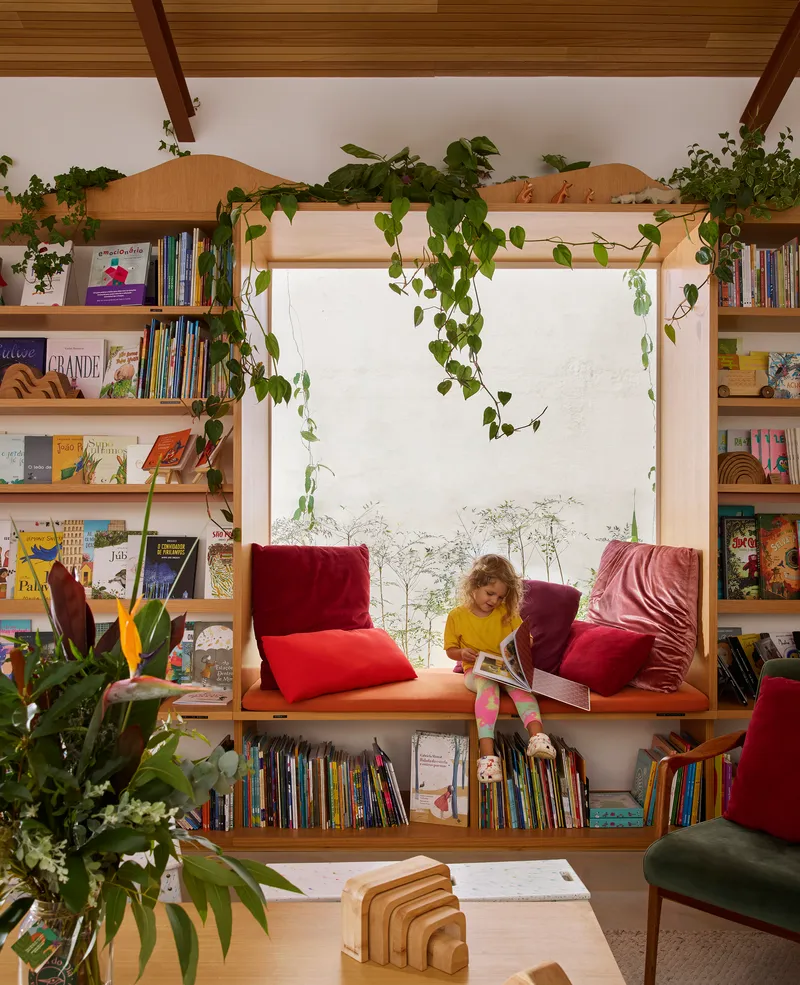 Built-in window seat with plywood bookshelves and cascading potted vines under exposed timber beams with child reading