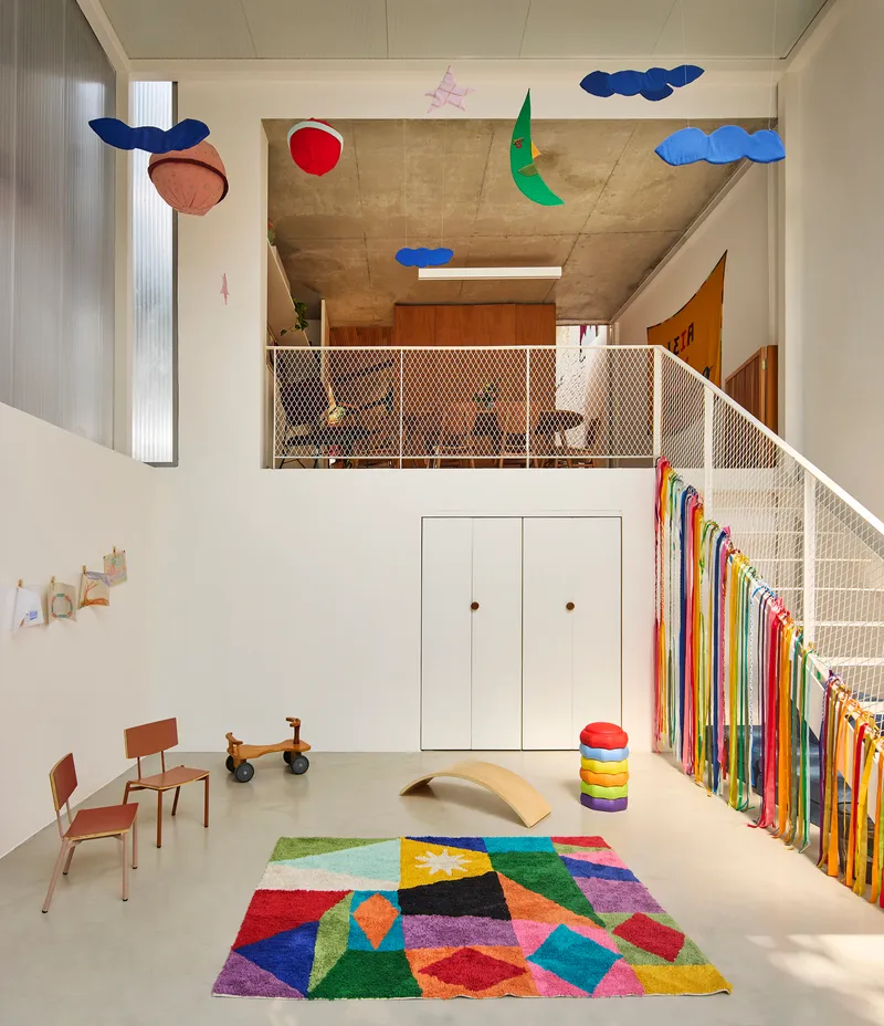 Double-height playroom with mesh-railed mezzanine, exposed concrete soffit, and suspended fabric mobiles above multicolored geometric rug