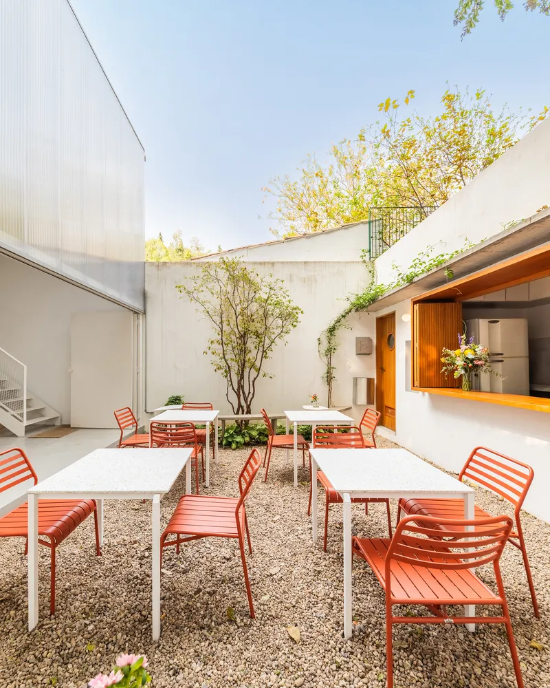Gravel courtyard with red metal seating and white tables framed by concrete walls and deciduous trees