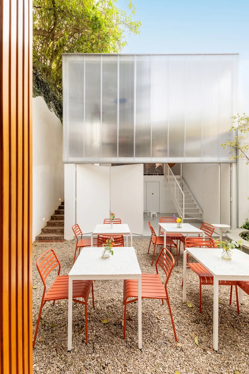 Translucent ribbed polycarbonate facade rising above the courtyard dining area with white metal stair visible behind