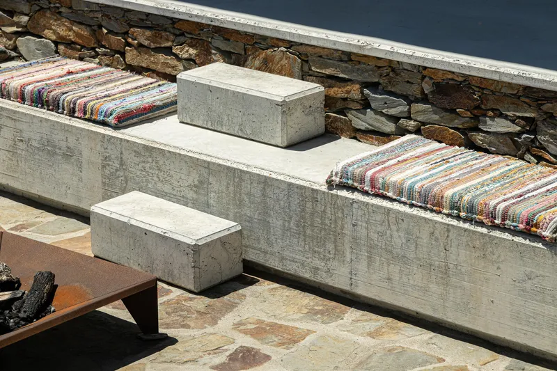 Board-formed concrete benches with striped cushions and stone side tables against a stacked stone wall in sunlight