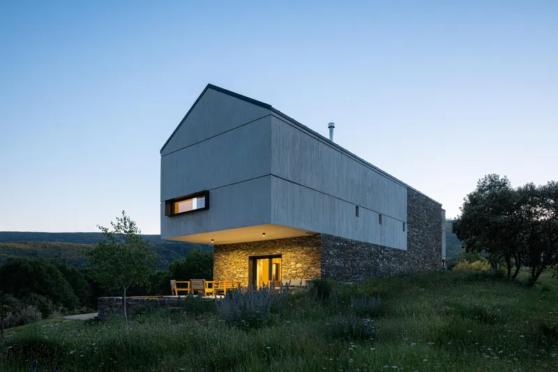 Cantilevered timber-clad volume over a stone base with illuminated entrance at dusk