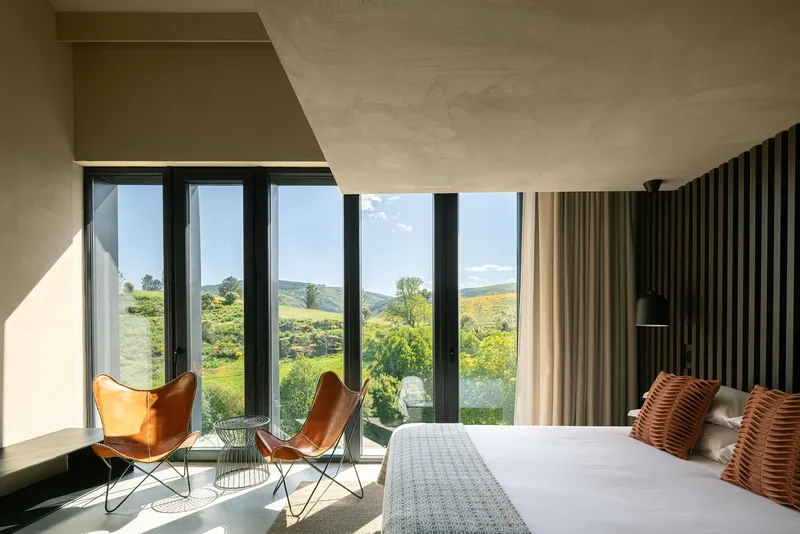 Bedroom with floor-to-ceiling windows framing green hillside view and vertical slat headboard