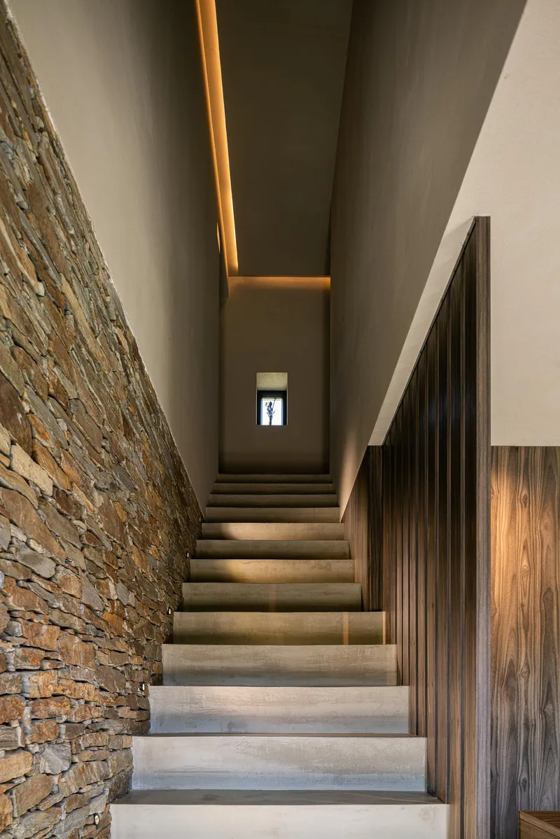 Concrete staircase with recessed ceiling light slot between layered stone wall and wood-grain paneling