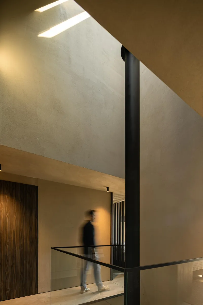 Double height hallway with angled skylight illuminating plaster walls and a figure crossing glass balcony