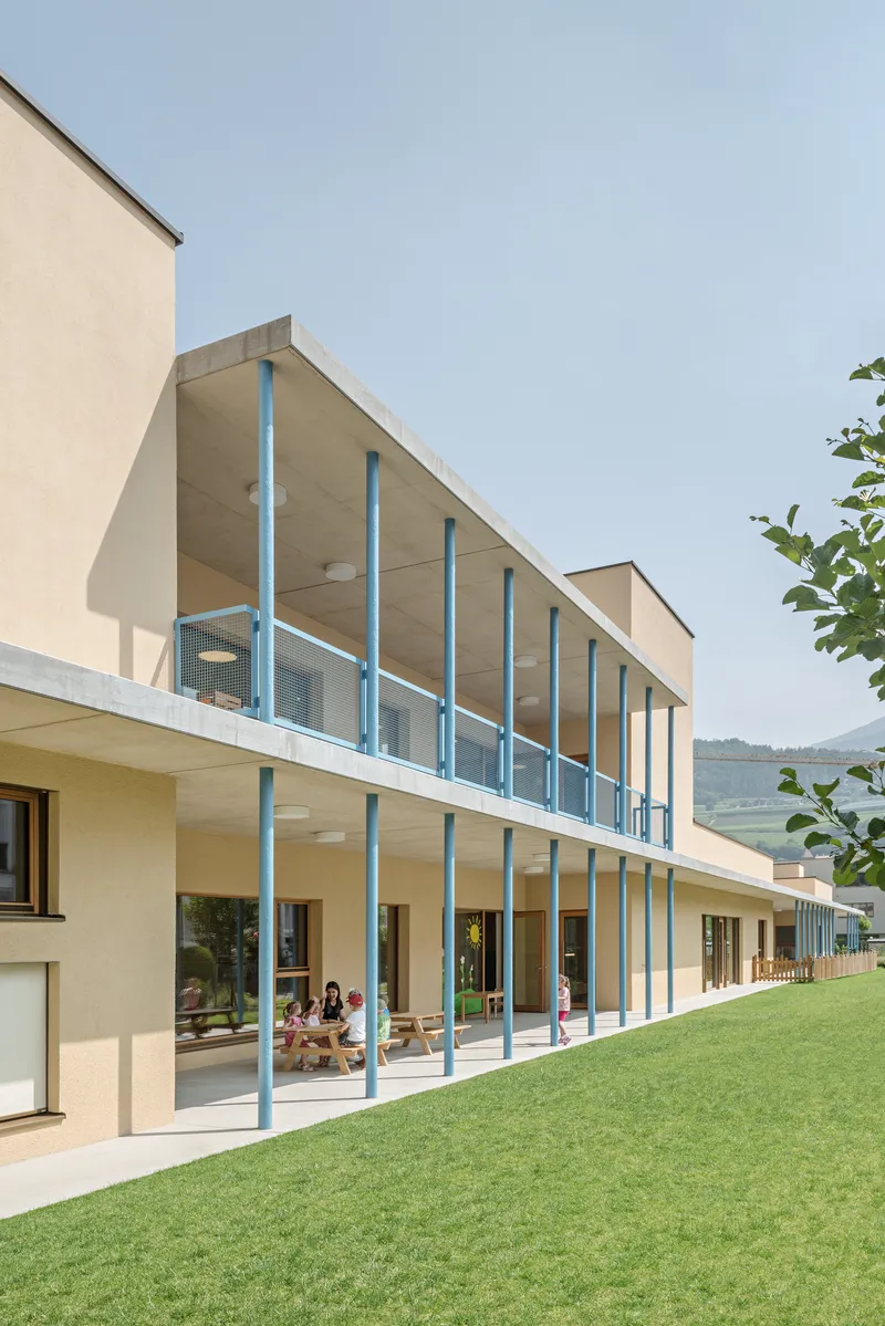 Two-story glass colonnade with slender blue supports facing a sloped lawn with children playing inside