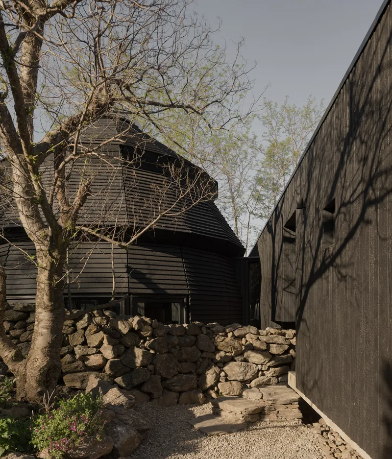 Gravel pathway between charred timber fence and stone wall leading toward the shingled dome volume