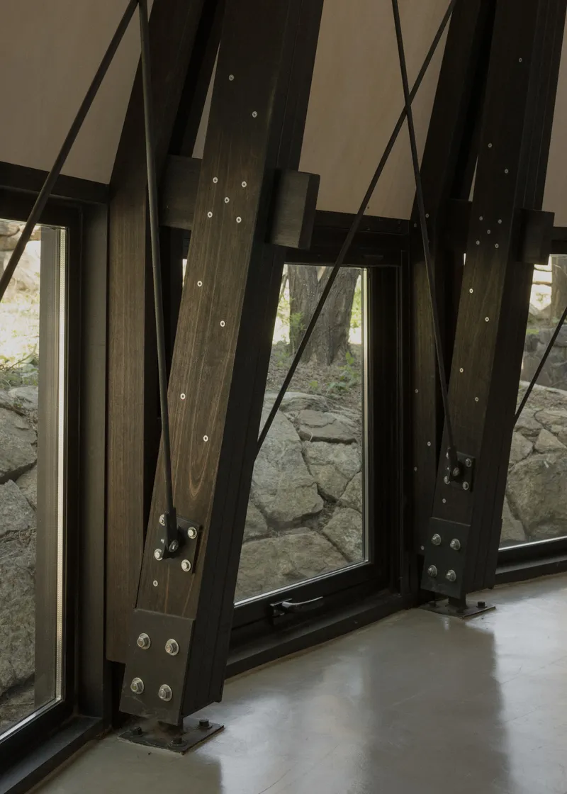 Diagonal timber bracing with bolted steel connections framing a window to a stone wall outside