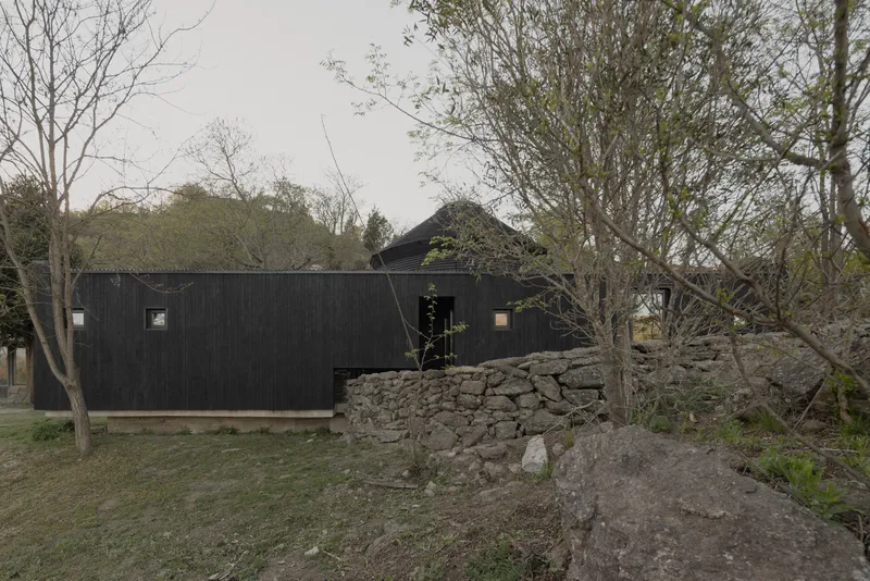 Long black timber box volume beside a dry-stacked stone retaining wall and bare trees
