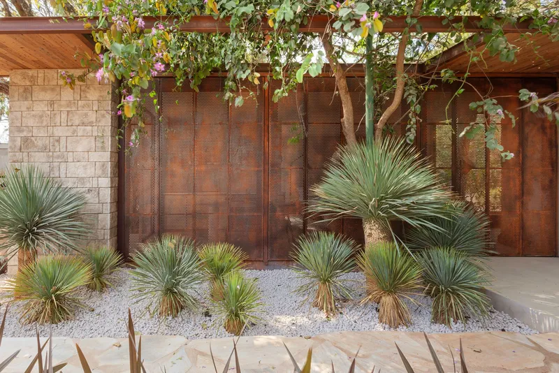 Perforated weathered metal screen wall with overhead timber pergola and planted yuccas in white gravel