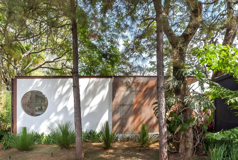 Exterior wall with circular window and weathered steel panels surrounded by tall trees and dappled sunlight