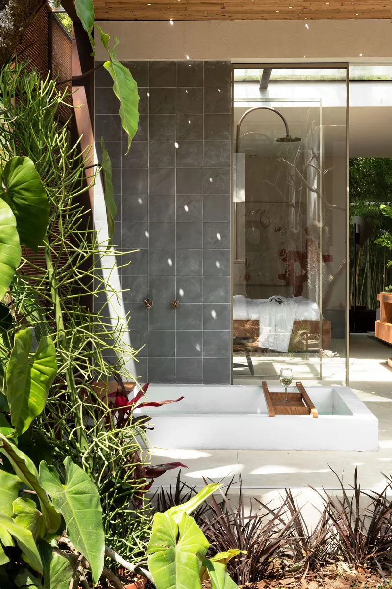 Outdoor shower with white basin beside grey tile wall and perforated metal screen among tropical plants