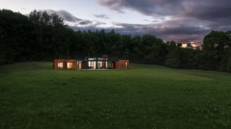Distant view of the horizontal timber and glass volume set in a meadow at dusk