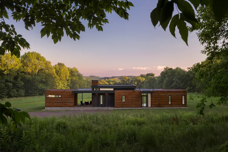 Front facade with horizontal wood cladding and dark glazed sections framed by overhanging tree branches at dusk