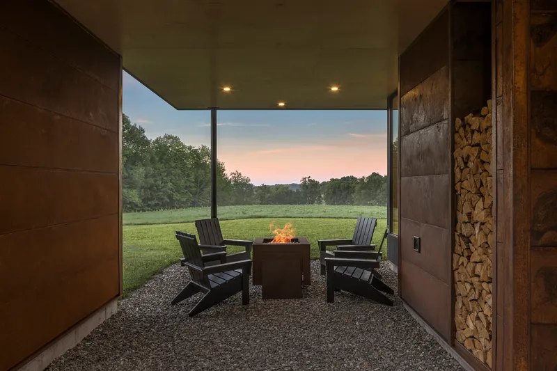 Covered outdoor terrace with fire pit and chairs on gravel looking toward open field at twilight