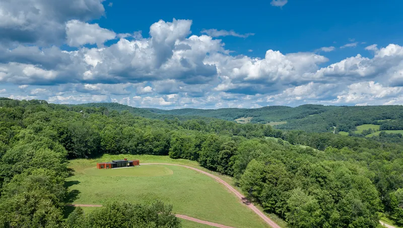 Aerial view of the residence on hilltop meadow surrounded by forested ridges under summer clouds