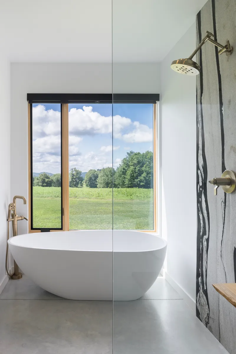 Freestanding white bathtub positioned before a tall window framing pastoral views under cloudy skies