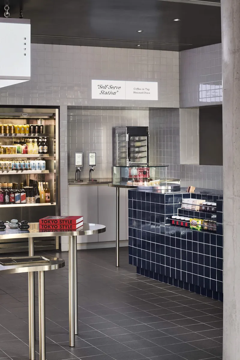 Self-serve station with navy tiled counter and illuminated shelving visible beyond glass vitrine and concrete column