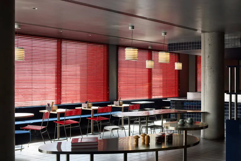 Dining area with red venetian blinds filtering daylight onto tables and exposed concrete columns