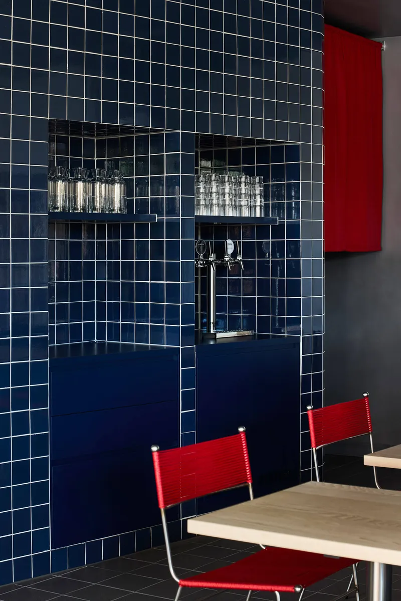 White-grouted blue tile wall with recessed shelving, chrome tap, and red canvas chairs at a table