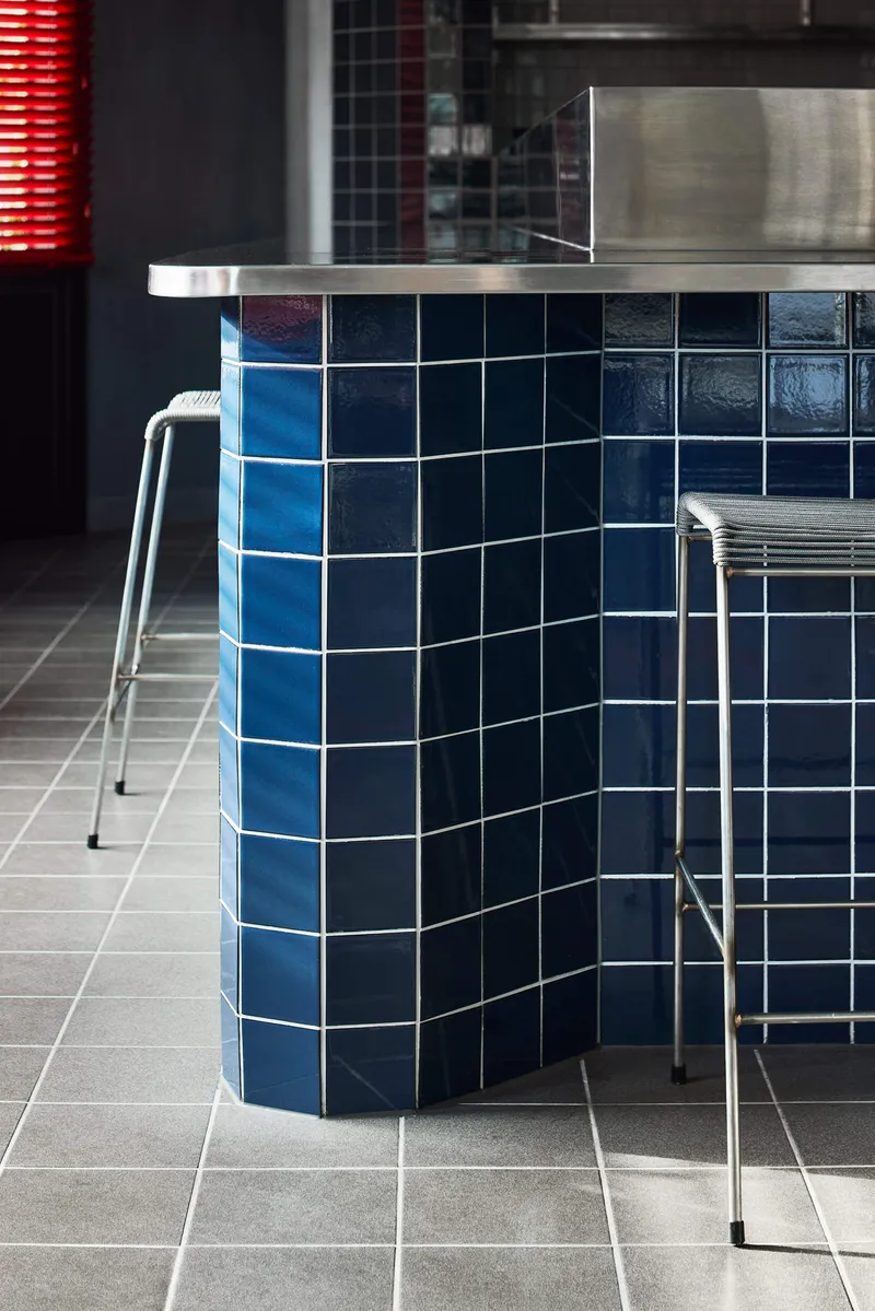 Curved blue tiled counter edge with stainless steel top and grey floor tile below a stool
