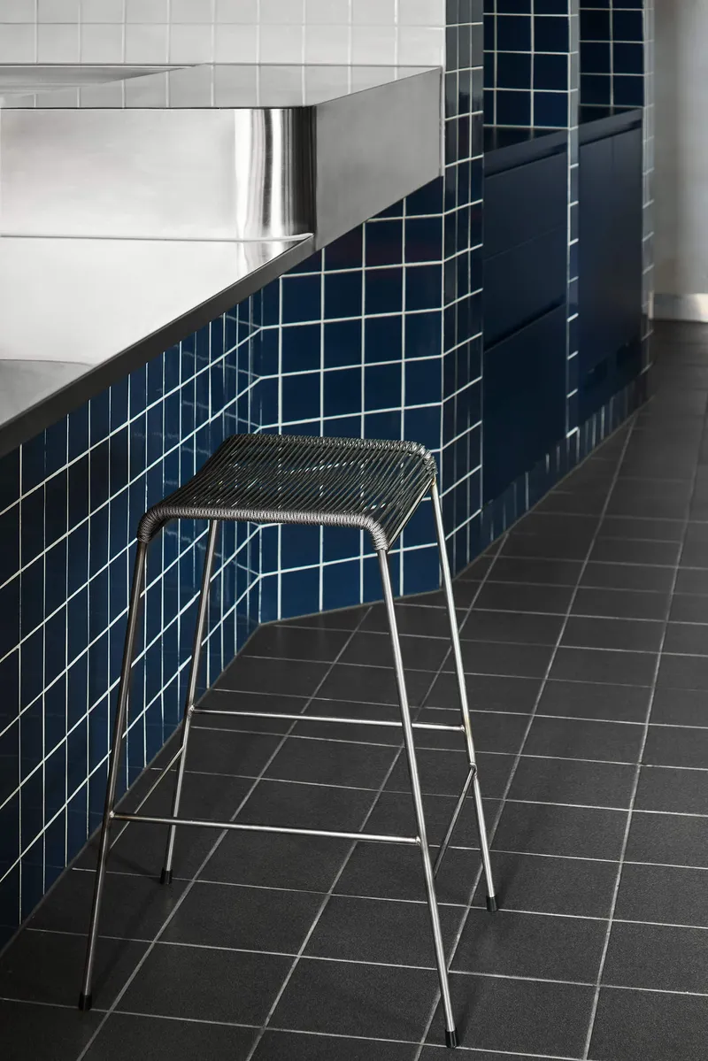 Wire mesh stool placed against the curved blue tile counter meeting white tiled wall and dark floor