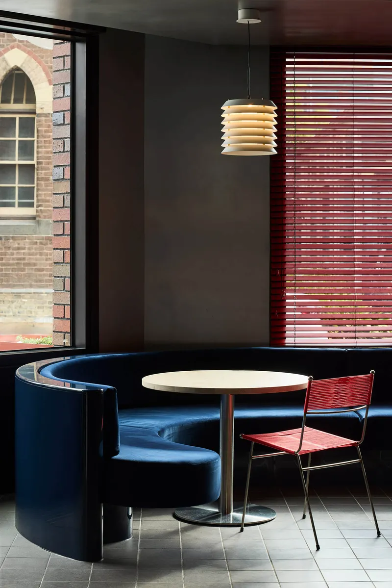 Curved blue banquette seating beside a round table with pendant lamp and red venetian blinds filtering light
