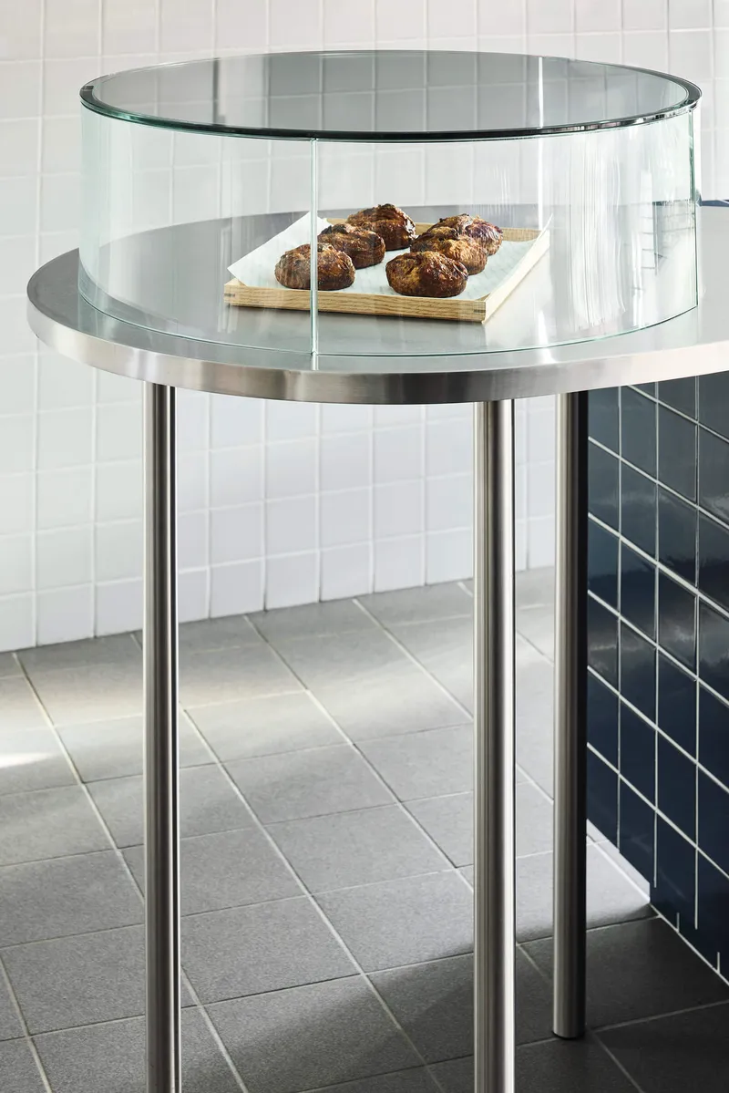 Cylindrical glass display case on chrome legs with white tiled wall behind