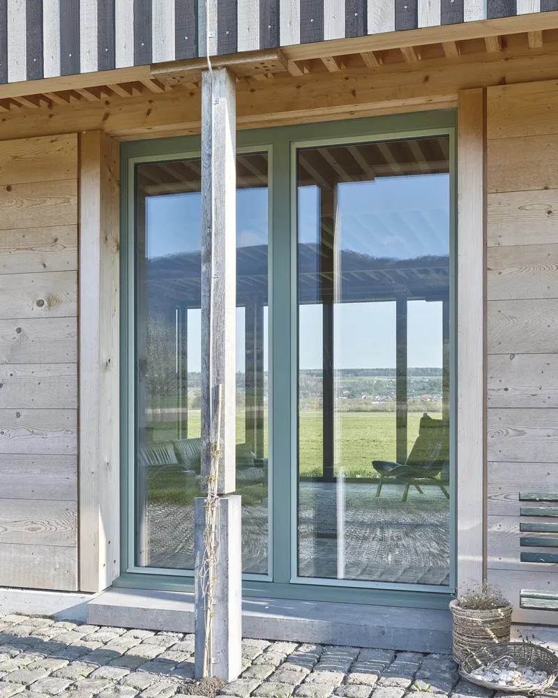 Green-framed glass doors in a timber and concrete facade reflecting the landscape under an exposed rafter ceiling