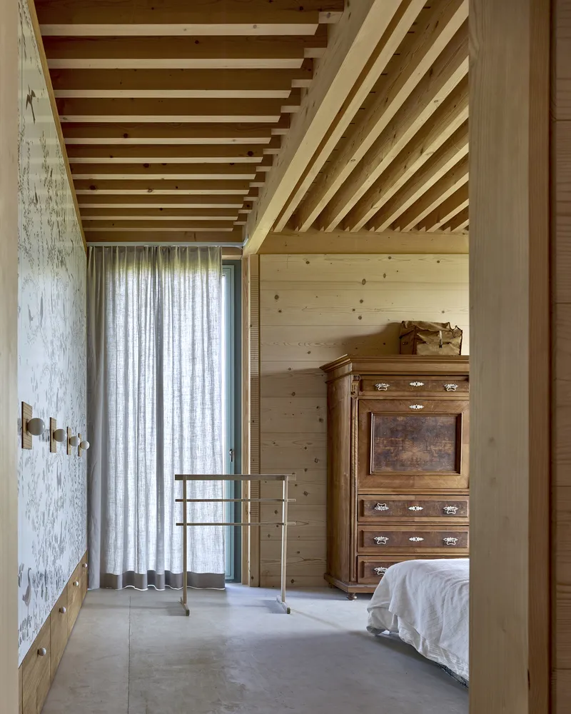 Bedroom with exposed timber ceiling joists and a pale curtain hanging beside a concrete polished floor