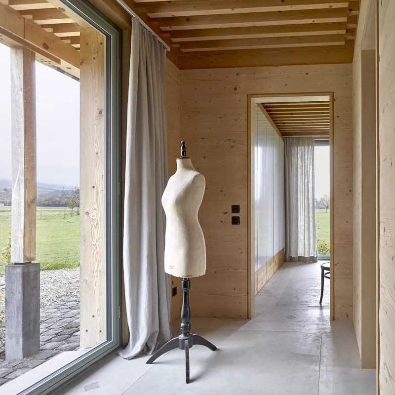 Interior corridor with exposed timber ceiling joists, floor-to-ceiling glazing, and a dressmaker's mannequin on polished concrete