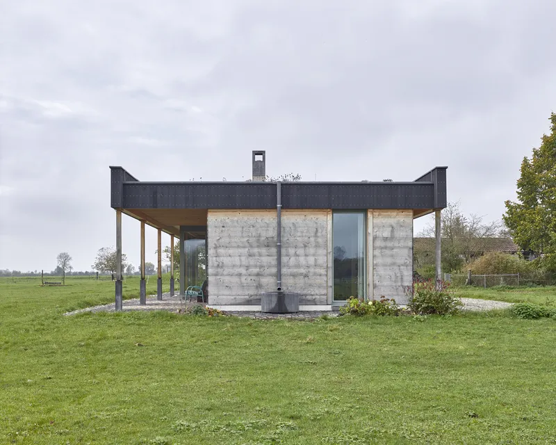 Board-formed concrete pavilion with metal-clad overhanging roof and central chimney in open meadow under overcast sky