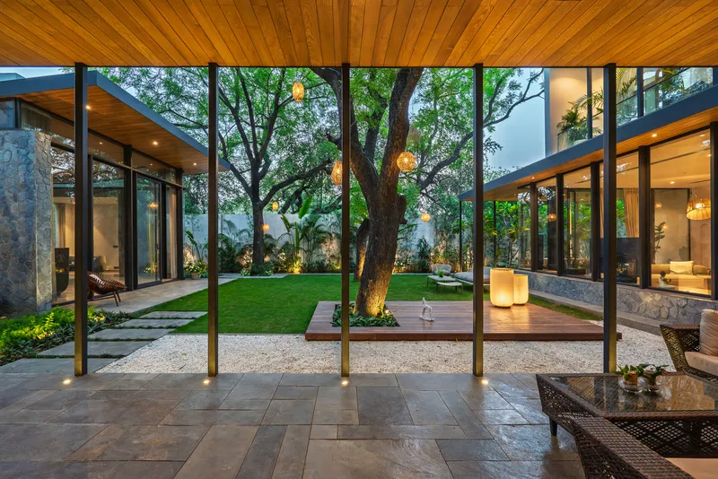 View through timber-framed glazing into a central courtyard with a mature tree and surrounding pavilions at dusk