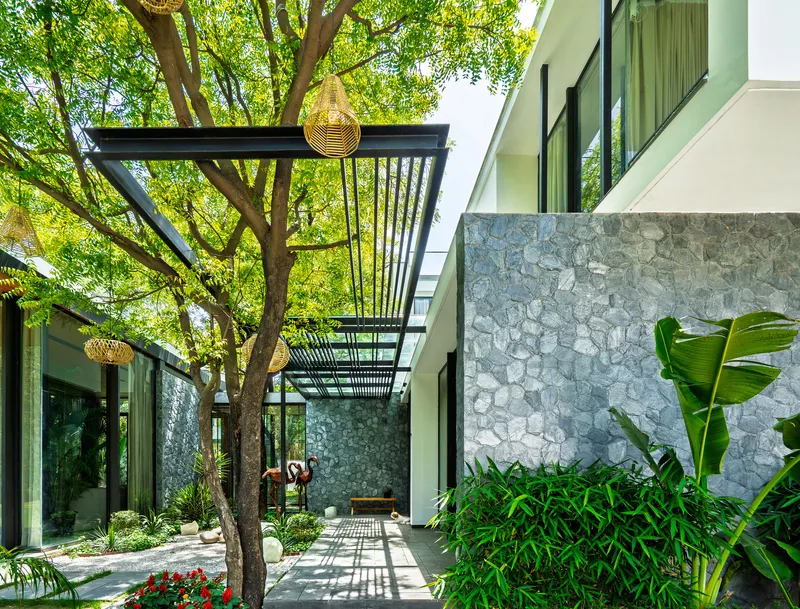 Covered walkway with steel pergola and woven pendant lights alongside stone wall and tropical planting