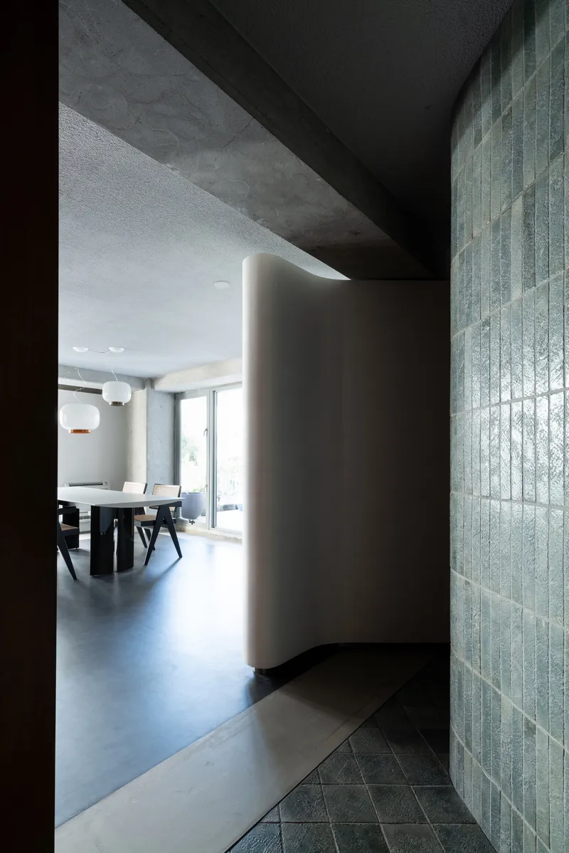 Hallway with curved white wall and vertical glass tile leading to dining area under exposed concrete ceiling