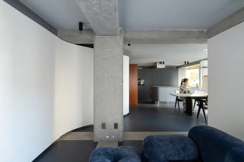 View through exposed concrete column toward open kitchen with person standing at counter in afternoon light