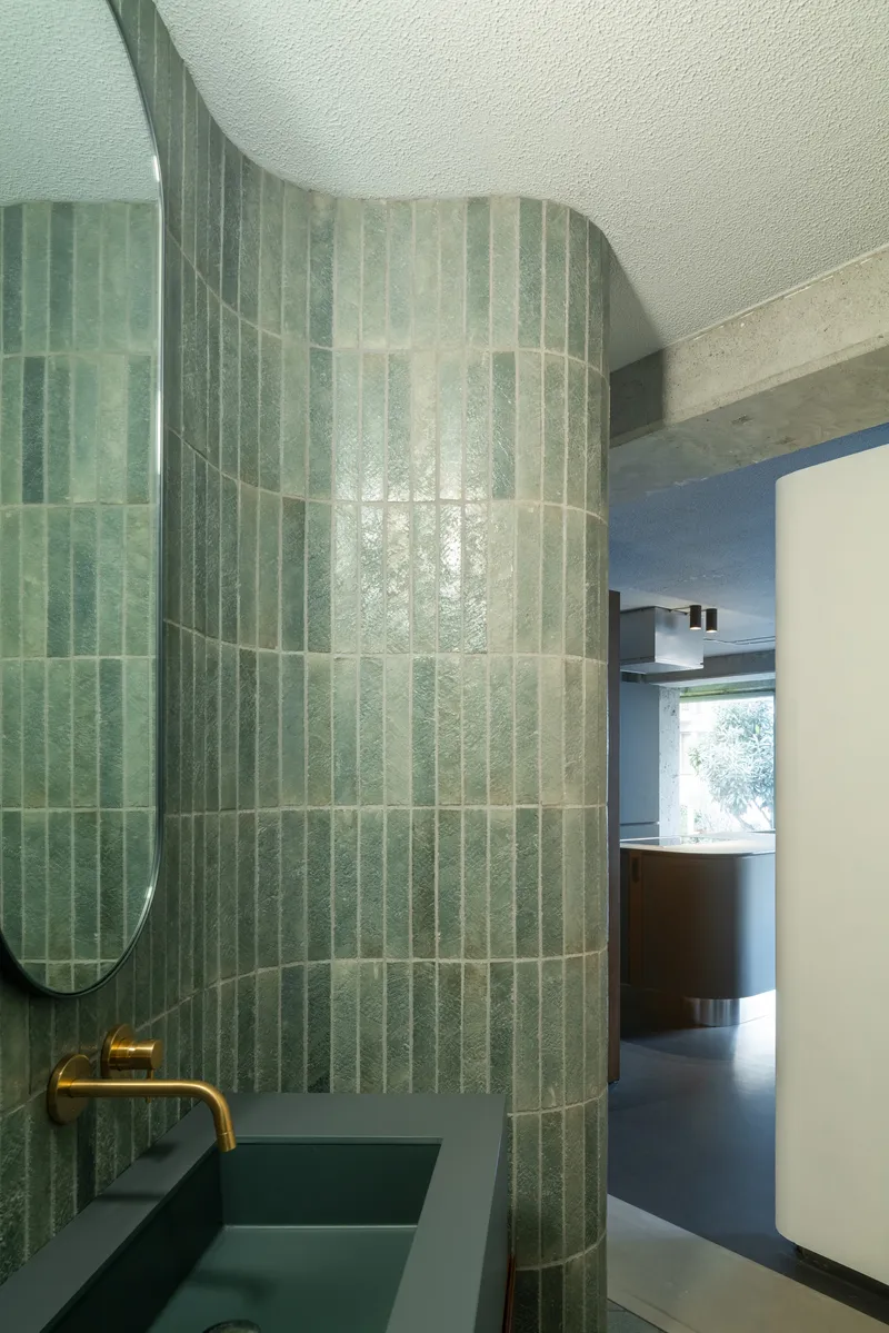 Curved green vertical tile wall in powder room with brass faucet and oval mirror reflecting interior