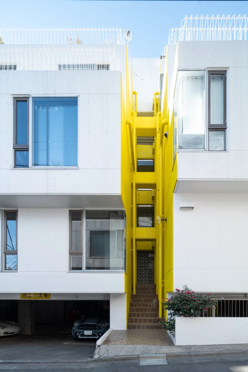 Yellow vertical stair shaft rising between white facade volumes with planted beds below under clear sky