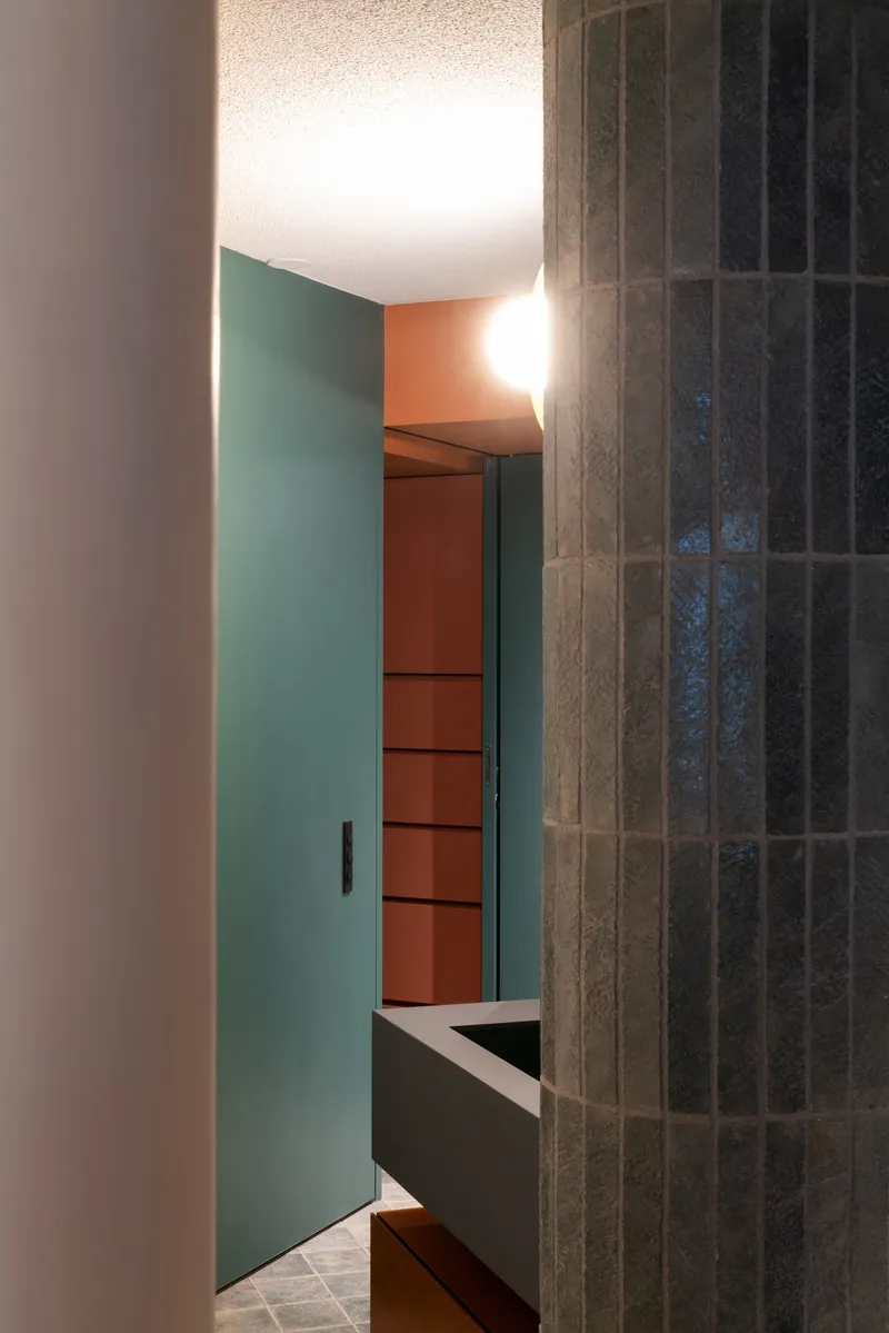 Doorway framed by teal wall panels and dark vertical tile leading to terracotta shelving beyond